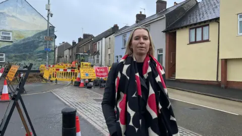 A woman wearing a black and pink colourful scarf is standing in front of the closed road. The yellow barriers are in the background behind her. Road closed signage is on displayed. There are a row of houses behind her. 