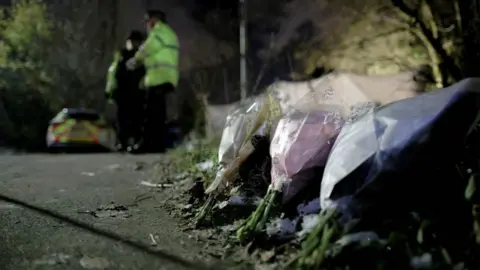 Three bunches of flowers are laid in a row on a path. Two police officers can be seen standing in the background.