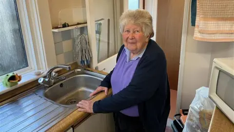 Phil Harrison/BBC An elderly woman with grey hair standing at her kitchen sink