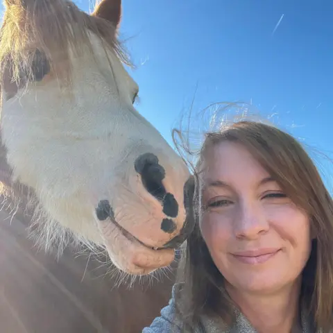 Philippa Davies A woman with long light brown hair smiling in a selfie with a brown and white pony