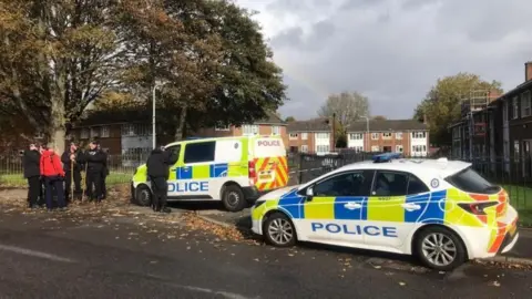 A police car and a police van block access to a residential side street. Several police officers wearing all black, some holding long sticks, stand on the left with a person wearing a red top. They are underneath a large tree. Terraced homes can be seen on the other side of the police vehicles.