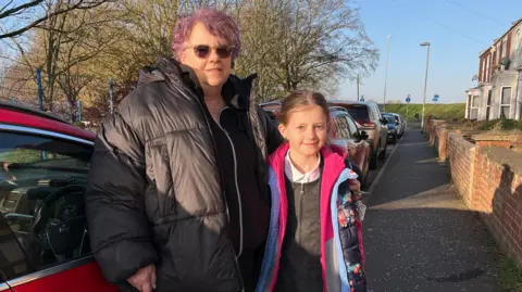 Fiona Cheethum and her granddaughter Emily stand outside a red car on the road outside the school. Fiona is wearing a black jacket and has light purple hair. Emily is 9 and wearing a pink patterned coat over her school uniform. 