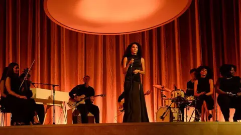 Getty Images Olivia Dean performs onstage during the 2026 MOBO Awards. She is wearing a black dress and singing into a microphone. There is a band behind sat behind her. 