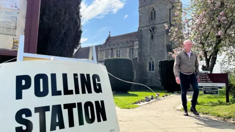 PA Media A large white sign reading “POLLING STATION” stands in the foreground outside a stone church with a clock tower and St George’s flag flying above it. A person walks along a paved path beside flowering trees and neatly trimmed hedges. Church noticeboards and directional signs are visible, with a bright blue sky and scattered clouds overhead.