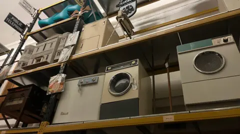 Large shelves holding washing machines from the 20th century as well as a model of a house and an ornate Barclays Bank sign.