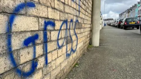 Newyddion S4C A brick wall with the word "grass" in dark blue graffiti paint. There is a pavement in front of it with a lamppost, and cars parked along the side.