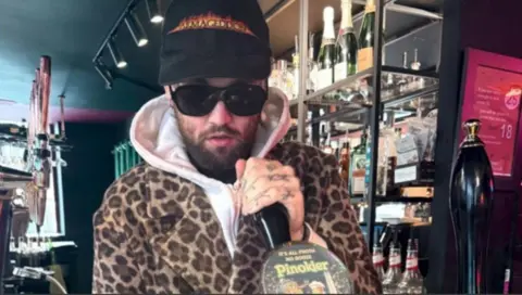 Mason Newman, who has a beard, stands behind the bar and pulls a pint. He is wearing shades and a leopard-print coat, as well as a black baseball cap. He is looking at the camera.
