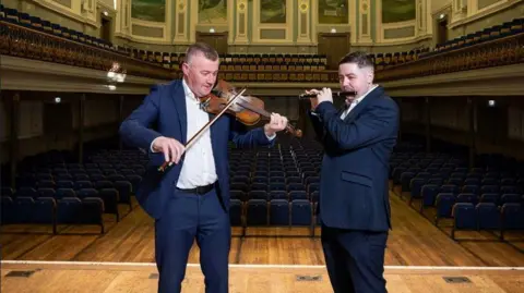 Belfast City Council A man on the left with fair hair in a blue suit and white shirt plays a fiddle, to his right is a man with dark hair and a blue suit and white shirt, playing a flute.