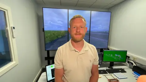 Mark Ansell/BBC Air traffic control trainee Liam Watts in the simulator room with the big screen, showing the virtual airport, in the background. He has blond hair and a beard and is wearing a cream polo shirt.