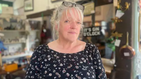 Simon Thake/BBC A woman with white hair tied back and black rimmed glasses at the top of her head. Behind her are an array of antiques