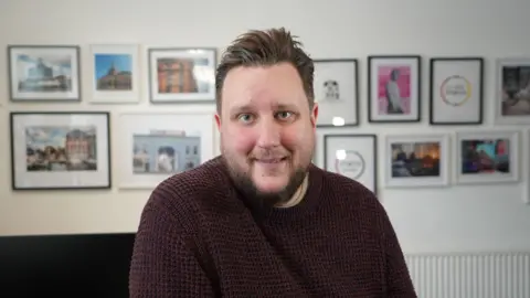 Qays Najm/BBC Lee Walker is leaning on a desk, with a gallery of big Ipswich events and landmarks on the wall in the background. He is wearing a burgundy jumper, and has a brown beard. 