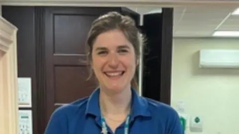 A woman wearing a blue NHS top and a lanyard is smiling at the camera. She has dark hair.