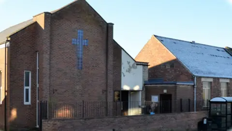 St Andrew's Methodist Church is a brown-brick building with a cross. It sits behind a black fence.