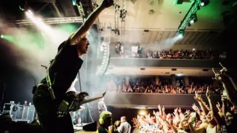The lead singer of McFly, Tom Fletcher, jumping on a stage with his right arm in the air. The picture is taken from close range at the front side of the stage and the audience can be seen in stalls and on balconies, raising their arms in response. A guitar is slung around the singer's shoulder on a strap. He is wearing black, has blonde hair and black glasses. Other band members can be dimly seen behind him on the stage. 