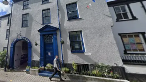 Google A tall beige building with several windows across three floors, each of which have blue frames. On the left there is an archway painted in blue, with the words 'The Brewery' painted on it in white. In the centre there is a blue door with an ornate glass panel above it. In the foreground, a man in a grey jacket is walking past, and there are black railings with green plants.