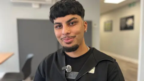 Emma Baugh/BBC Ali, a boy with short black hair, wearing a black Shirt, smiling for camera.