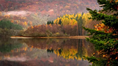 A photograph of a loch during autumn. Trees with red, orange and yellow leaves cover a hillside and reflect onto the calm water.