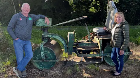 David wears a grey top with the Battisford & District club logo on it, while Julia wears a green jacket and denim jeans. They are standing at either end of a green old cricket pitch roller, in front of some trees.