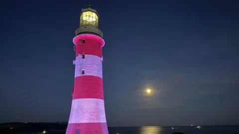 BBC A picture of Smeaton's Tower lit up at night on Plymouth Hoe with the moon reflecting on the water in the background.
