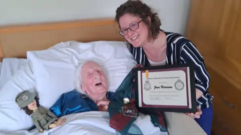 A picture of Joan lying in bed being presented a certificate. She is next to a toy and another woman who is presenting the certificate.