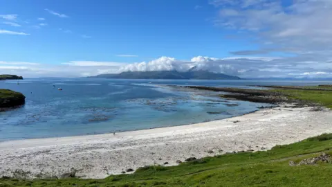Gav Burnage A sweeping coastal landscape featuring a white sandy beach, crystal-clear blue water, and distant mountains partly draped in clouds under a bright sky.
