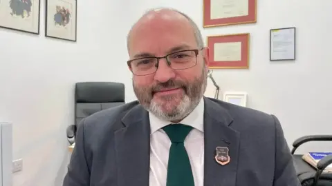 Simon Collins, wearing a grey suit with a white shirt and bottle green tie, sits in an office with framed certificates on the wall and two leather chairs in the background.