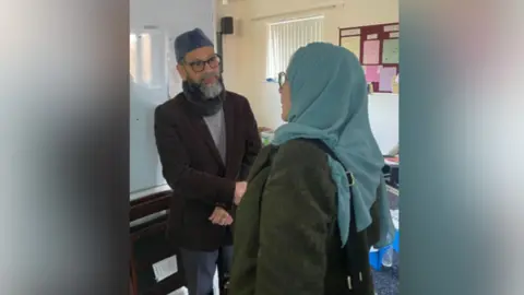 Office of the police, fire and crime commissioner A man wearing a blue skull cap, with glasses and a grey beard, is shaking hands with a woman wearing a blue scarf over her head and a dark green coat. They are in a training room with white walls and a noticeboard.