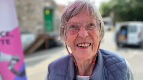 A lady with grey hair and glasses. She's wearing a blue ribbed gillet.