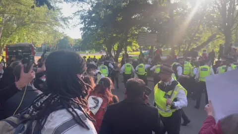 PA Media A group of police officers in high vis jackets at the scene of a Palestine Action protest outside Woolwich Crown Court. A large group of protesters can be seen on either side of the road.