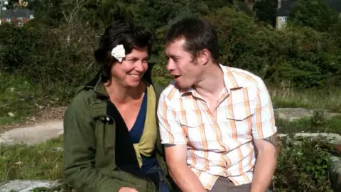 Cara Lavan Cara is sat with Jake Coe on a rock. Cara is smiling wearing a green jacket and white flower in her hair. Jake is sat looking at her. He is wearing a white shirt with orange stripes going down it.
