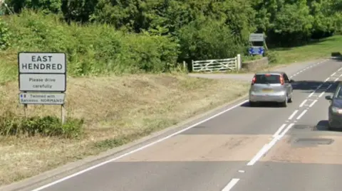 The A417 Reading Road in East Hendred. It is a wide single lane road. There is a white sign next to the street that reads 'East Hendred'.