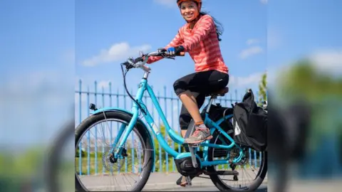 A woman on a turquoise bicycle, smiling to camera, with blue iron railings and greenery in the background. She is wearing a pink striped top, black knee length leggings and trainers.