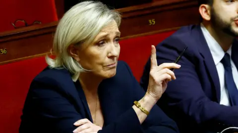 Marine Le Pen, who has a blonde bob and wears a blue blazer with a gold bangle, holds a hand up at the National Assembly in Paris