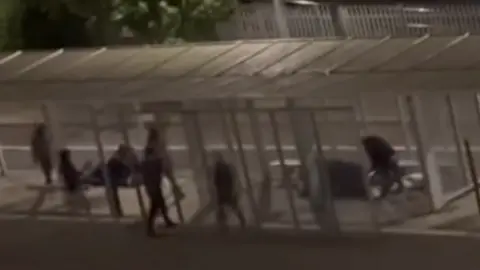 A grainy still from CCTV showing a group of people in dark clothes in a bus stop