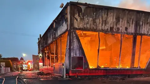Orange flames engulf a building. The windows of the building have been smashed and black suit covers the outside walls. On the left of the image a fire truck and crew can be seen. There is a large hose on the ground. 