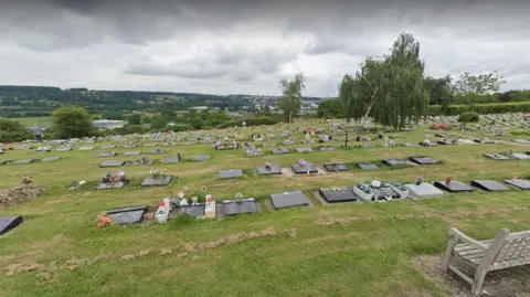 Google A view looking down the hill at South Bristol Cemetery, with hundreds of grave sites and headstones visible in the grass. There are trees planted at the cemetery, and it has a landscape view of fields and hedgerows.