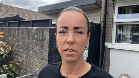 Shows a woman with dark hair up in a bun, wearing hooped earrings and a black t-shirt standing in front of a grey brick wall