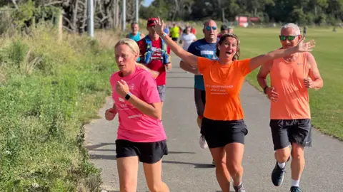 A runner smiles at the camera with her arms spread wide as she runs with a group of people