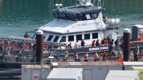 Border force boat with migrants walking on board wearing life jackets 