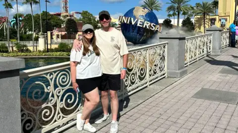 Melissa Rutter Daniel King A woman with a white t-shirt, black shorts, white trainers, a grey baseball cap and sunglasses standing next to a man wearing a cream t-shirt, black shorts, white trainers, a black baseball cap and sunglasses against white railings, with the Universal Orlando resort in Florida pictured the background.