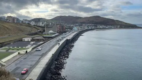 McGarrigle Architects An aerial view of Peel promenade, a long stretch of grey tarmac leads to a built-up area with multicoloured buildings. You can see rolling hills in the background.