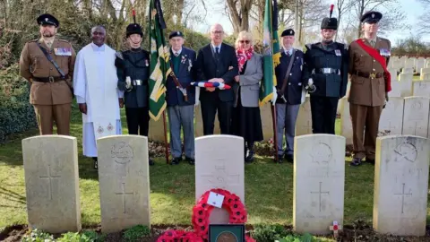 Crown Copyright Civilians, a chaplain and military personnel in uniform stand in a row behind a line of headstones in a cemetery. Poppy wreaths and medals stand in front of one of the graves. It is a sunny day and trees can be seen behind