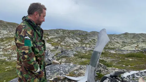 Chris Bullock Chris Bullock is looking down at a memorial to Group Captain Wilfred Surplice. Chris is standing sideways, wearing a green and brown camouflage jacket, and his hands are in his pockets. The memorial is made from part of a World War Two plane propeller. Behind him is a scene of scrubby grass and white and grey rocks.
