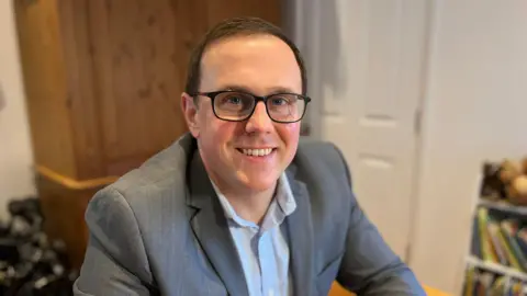 Ted Powell has glasses and sits at a dining room table wearing a blue shirt and a grey jacket. There are books on a shelf behind him.