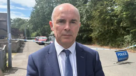 Alex Easton stands in front of a police cordon, blocking off a road beside some houses. On the right are tall, green trees. Mr Easton is looking directly at the camera with a neutral expression on his face. He has short, balding white hair and is wearing a blue suit jacket, a white collared shirt and a blue and white patterned tie.