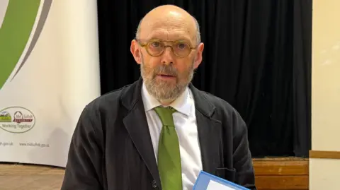 Louise Parker/BBC A man who is bald with a thick grey beard looks at the camera. He wears glasses along with a black jacket, white shirt and green tie. 