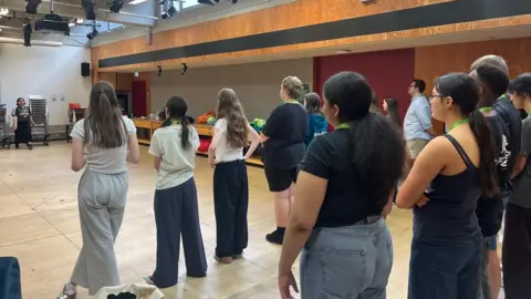 Abbey Grange Academy School students in a rehearsal room. They are standing in rows on a wooden floor, watching a teacher at the other end of the room.