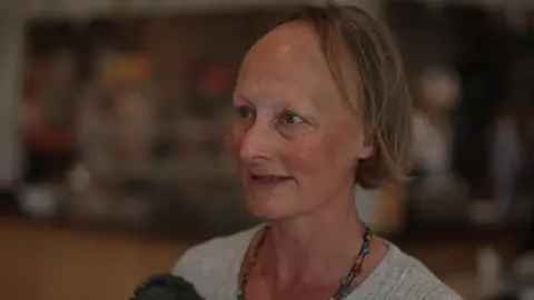 BBC Jenny Tagney with brown hair that stop just before her shoulders. She is wearing a grey cardigan with a beaded necklace.