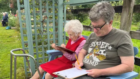 An elderly lady wearing a red pinafore dress and pink jumper draws leaning on a clipboard. She is sitting on a blue wooden bench in a green arbour, next to a middle aged man with thick dark grey hair and black framed glasses, who is also drawing with a pencil onto paper on a clipboard. He is wearing a dark green t-shirt with a logo that reads super champ and has a cartoon dog and a football image