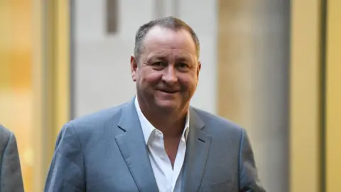 Mike Ashley smiling wearing an open-collar white shirt and a grey suit jacket